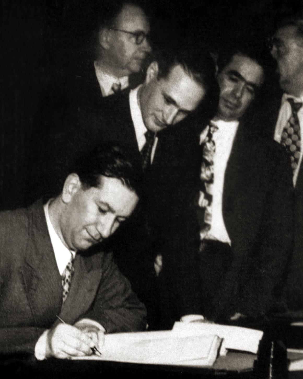 A black and white photo showing a man signing a document and other men standing behind him.