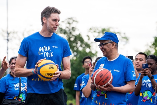 A tall man stands next to an average height man, both holding basketballs and wearing blue Walk the Talk t-shirts.