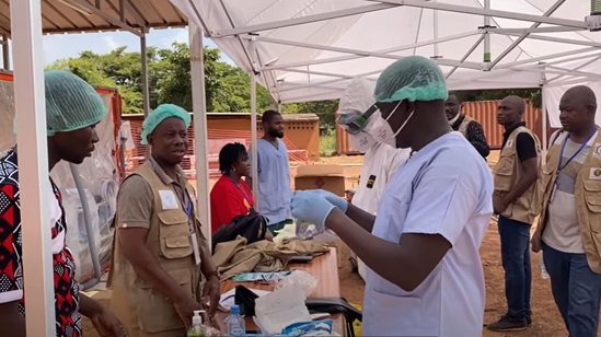 A group of people gathered under a tent roofing, some wearing full or partial personal protective equipment, others are wearing official vests and lanyards.