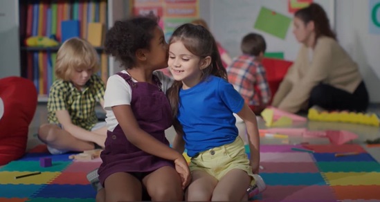 Two little girls sitting on the floor with other children in the background