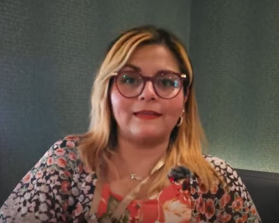 Video snapshot of a woman wearing glasses, speaking into the camera.