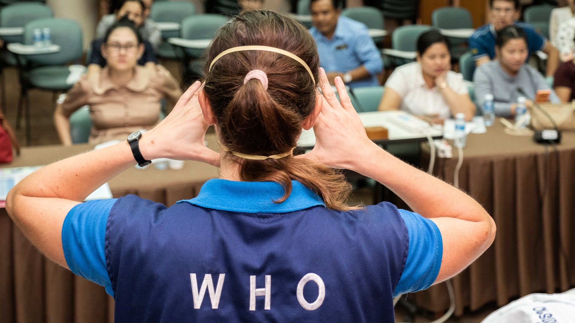 Photo 3 A WHO consultant wearing blue is standing in front of a class with their back to the camera. Their hands are raised, adjusting their facemask.