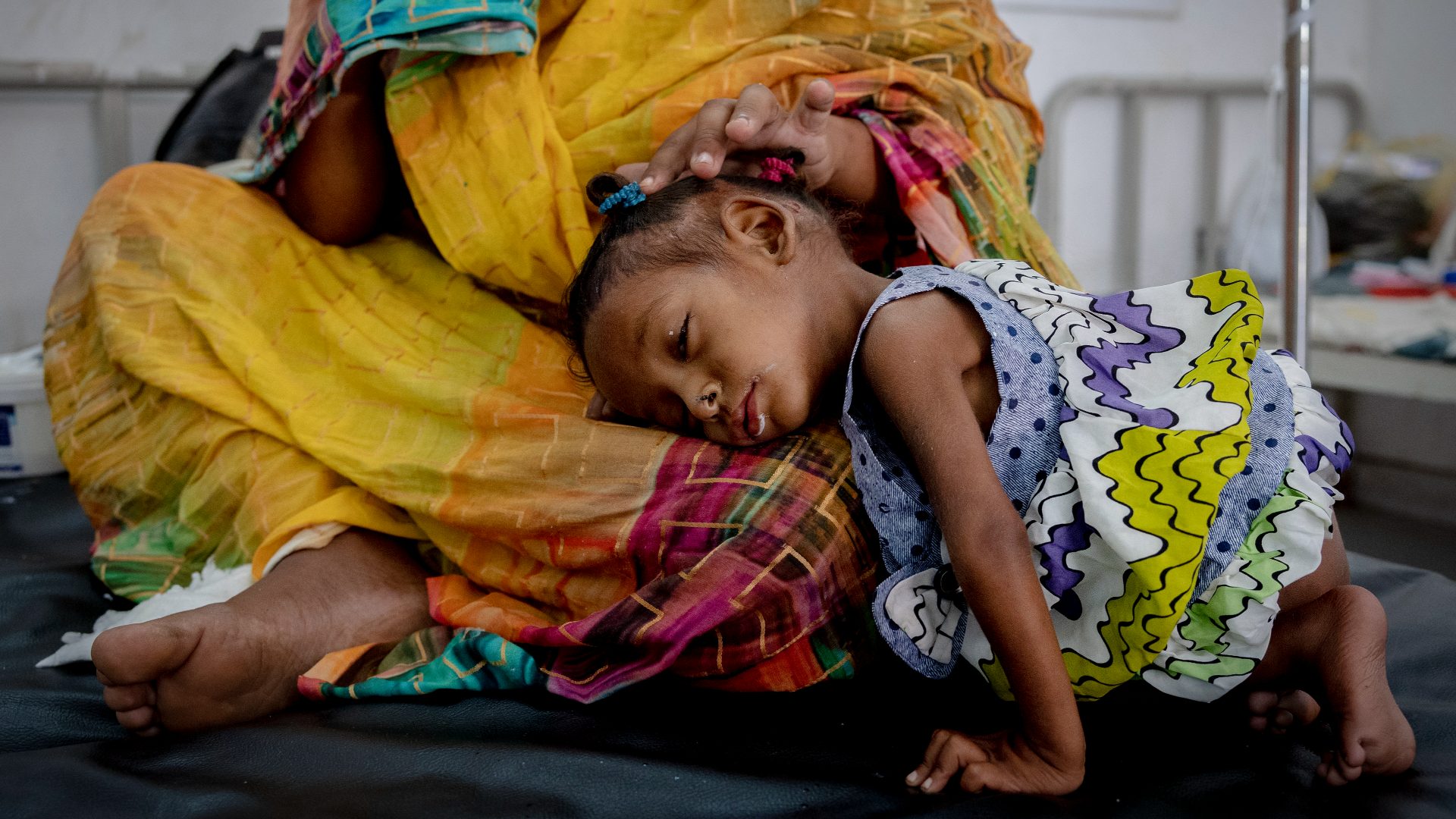 A young girl is resting with her head in her mother's lap. They are on a bed in a hospital setting.