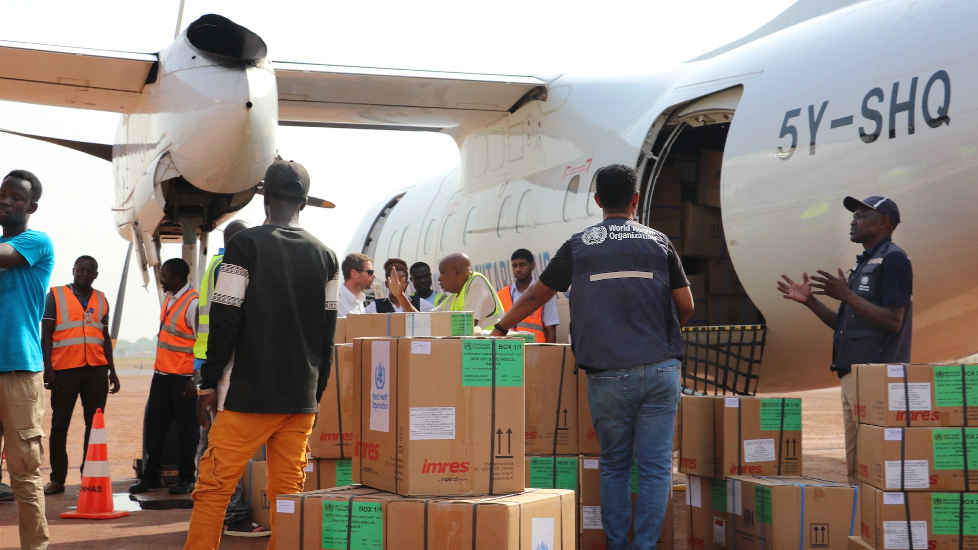 WHO officers and other people are standing next to an airplane with a large number of boxes on the ground.