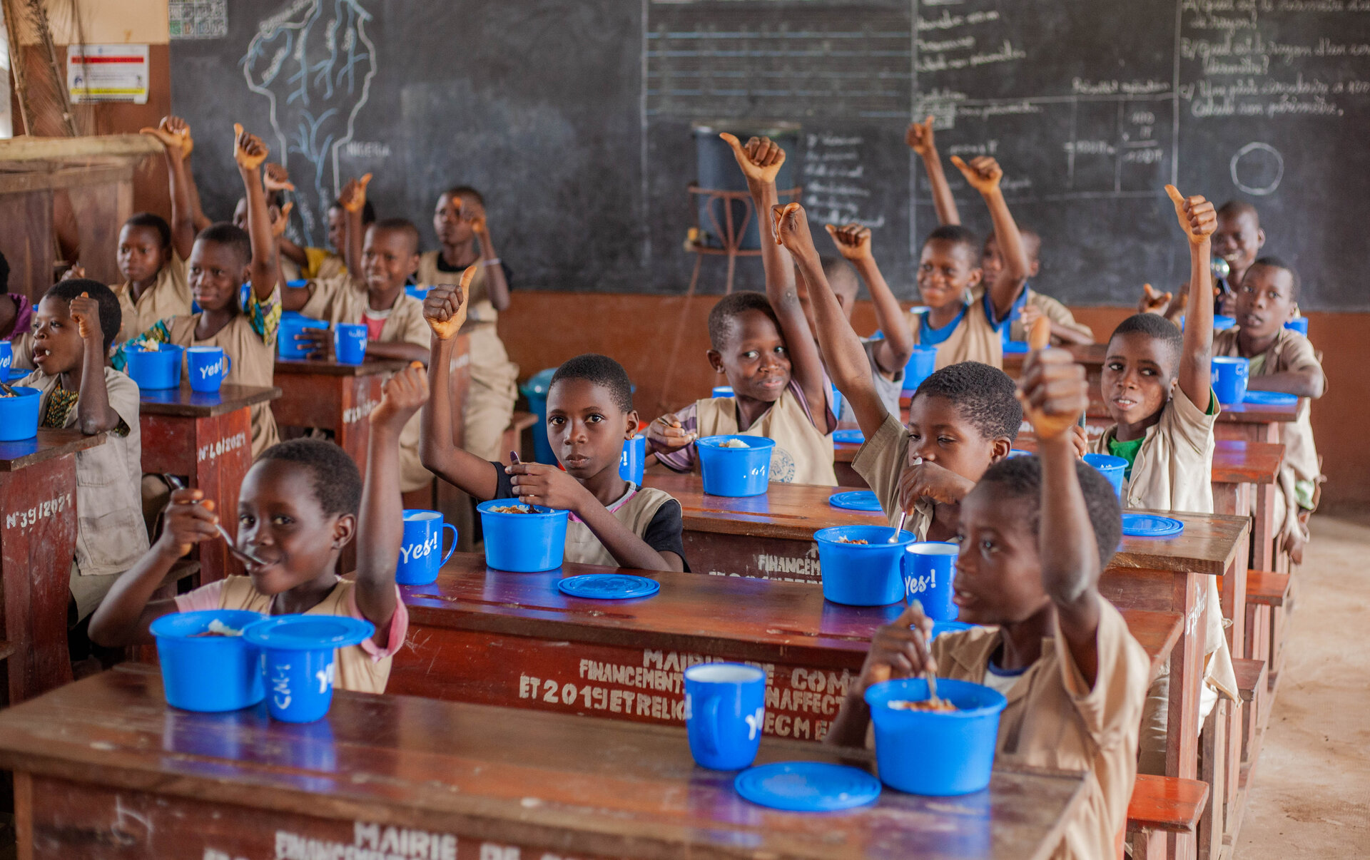 Children in a school have lunch while raising their thumbs to the air