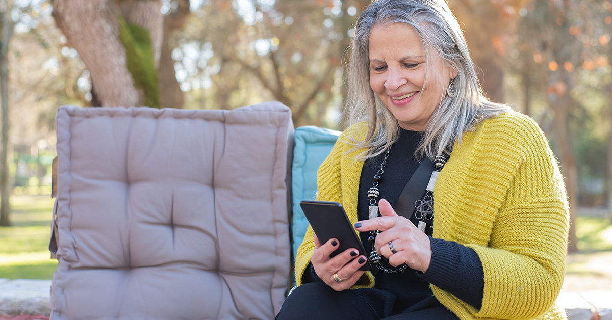 An older woman sitting on a bench outside, smiling while she scrolls through a smartphone.