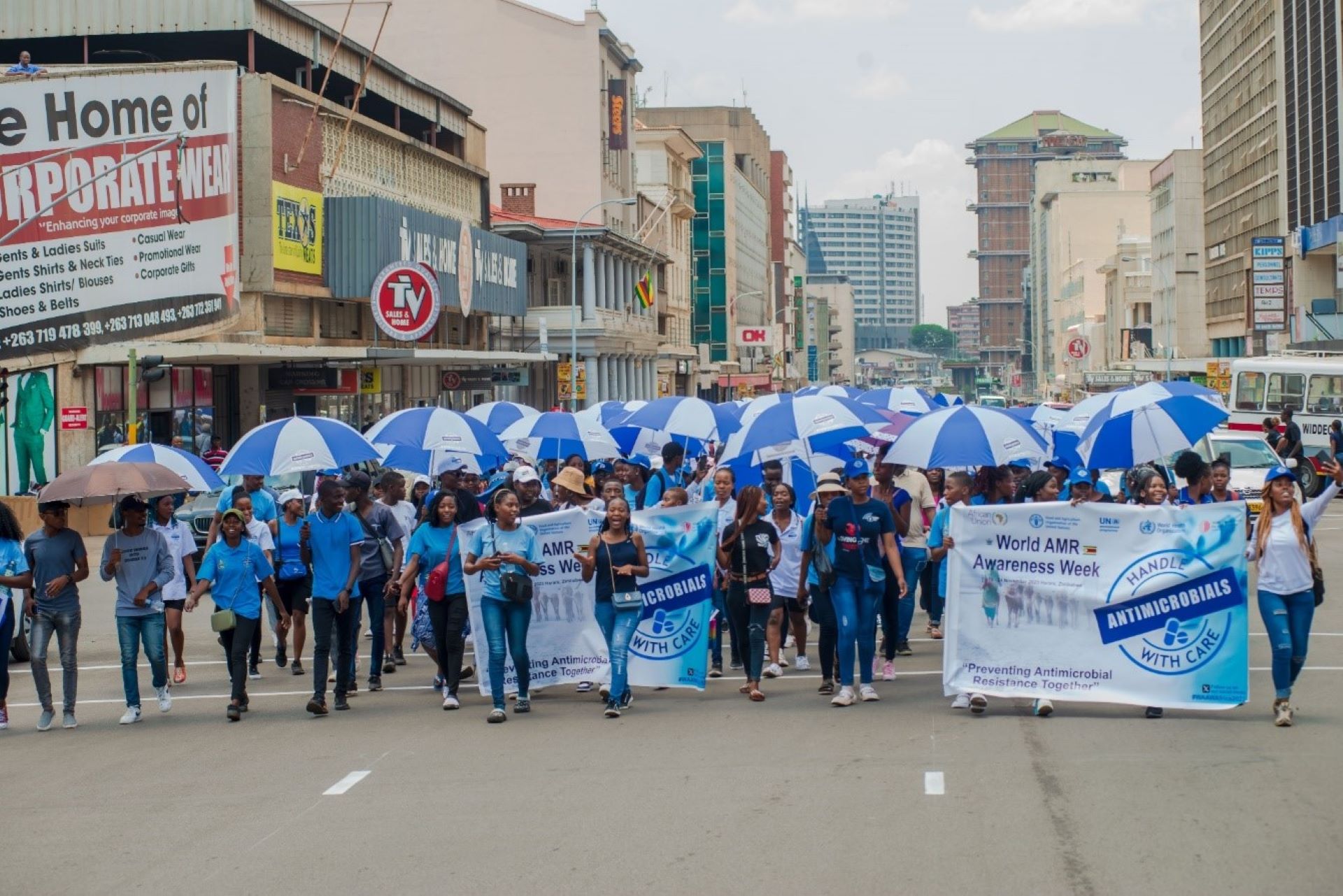 WAAW street campaign Group of people walking together on the street dressed in blue t-shirts, holding blue umbrellas and the poster with words: "World AMR Awaness Week: Handle antimicrobials with care"