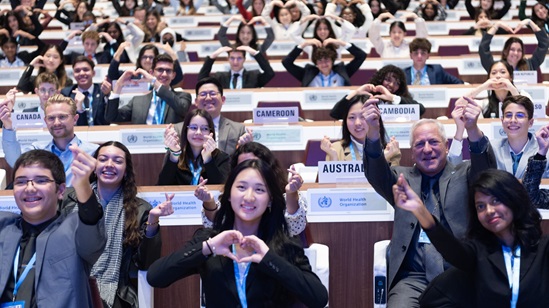 Young people sitting in a large conference room making heart shapes and other gestures as they smile and celebrate.
