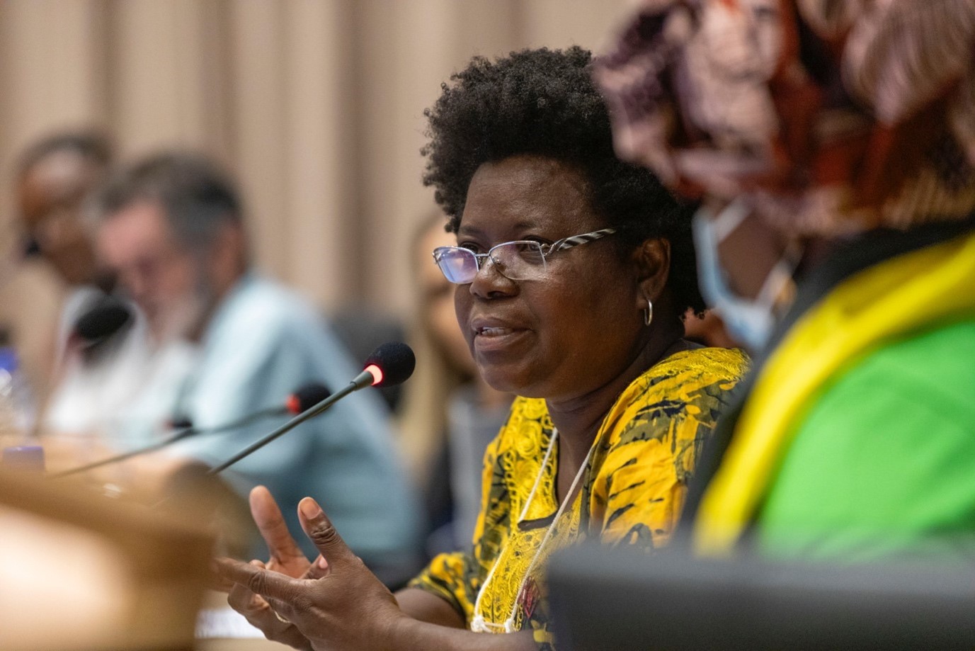 A woman wearing glasses and a yellow shirt is addressing attendees during a meeting.
