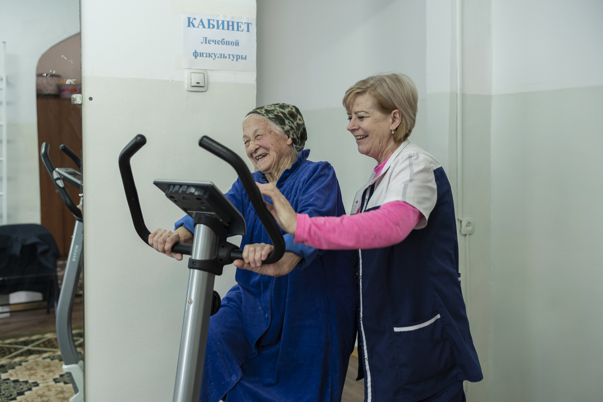 Elderly woman exercising on an indoor bicycle helped by a physical therapist