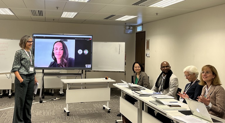 A trainer standing in front of a desk with 4 people seated, a trainer is on a screen in the background
