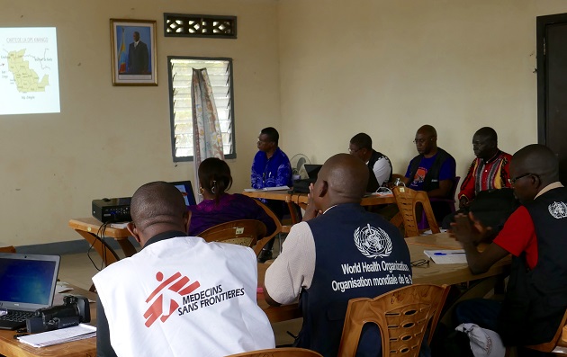 People wearing WHO and Médecins Sans Frontières vests meet in a room and use a projector to discuss operations.