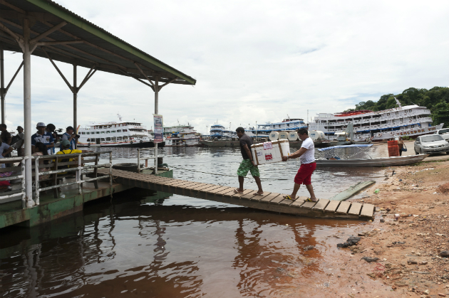 Two men carry a cooler box of vaccines over a wooden bridge into a boat, in Brazil