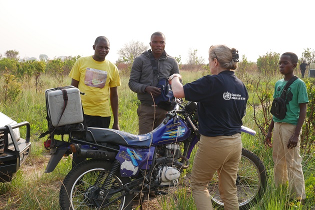 A woman wearing a WHO vest speaks to two men in front of a motorcycle in a rural area.