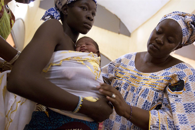 A new mother holds her baby wrapped around her, as a midwife helps