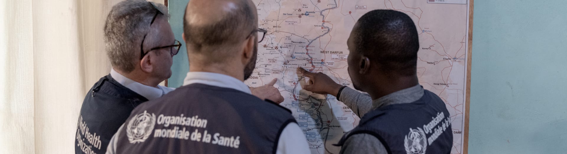 Three men wearing navy blue WHO vests pointing and looking at a map on the wall.