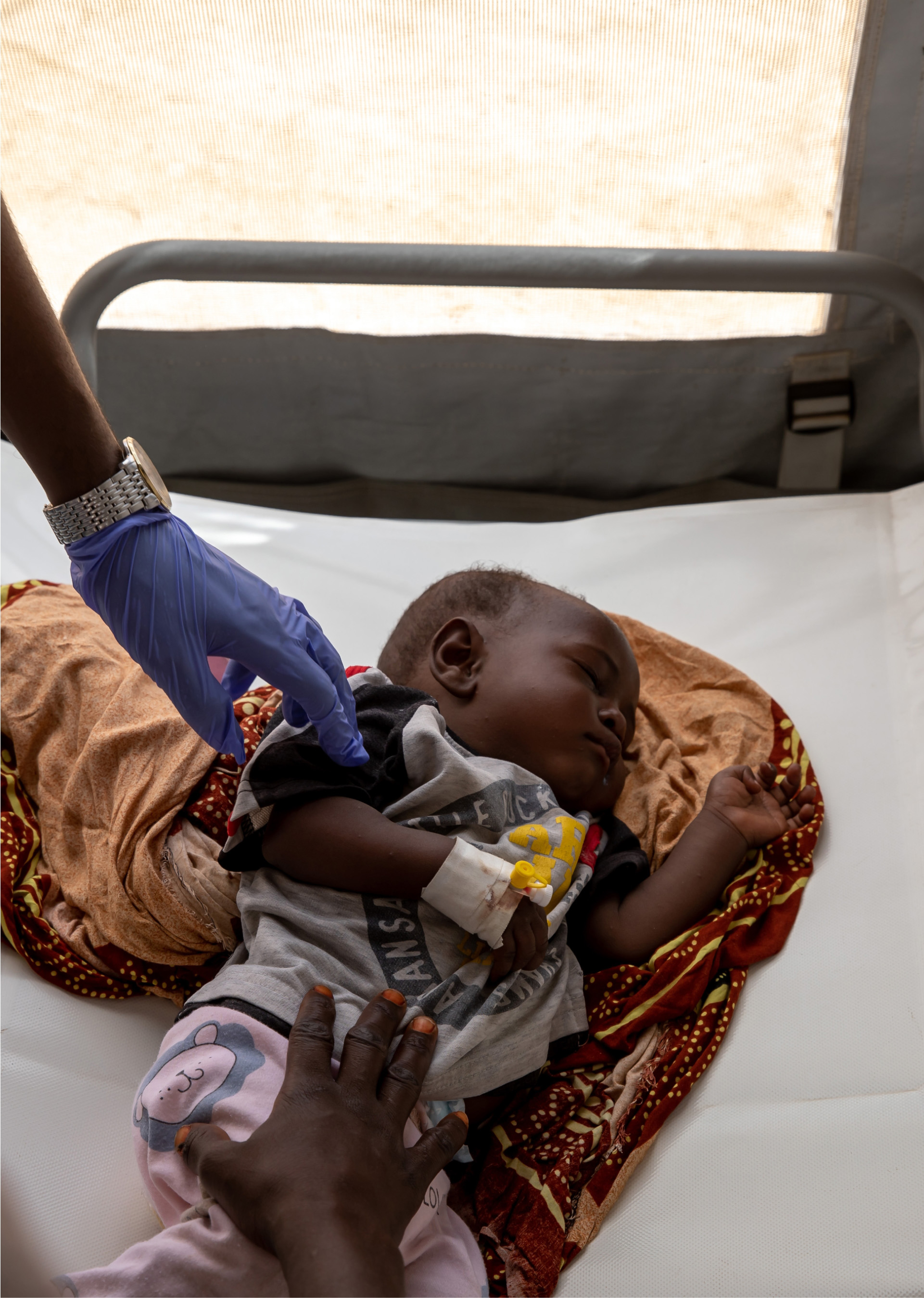 WHE 2025 Appeals listing Somalia A sleeping baby lying on a cot with an adult hand wearing a surgical glove touching its arm and another adult hand holding its lower body.