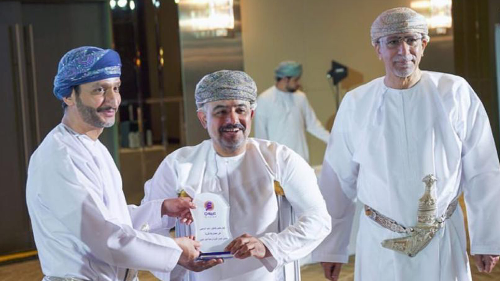 Three men wearing turbans, whereas one is receiving an award plaque.