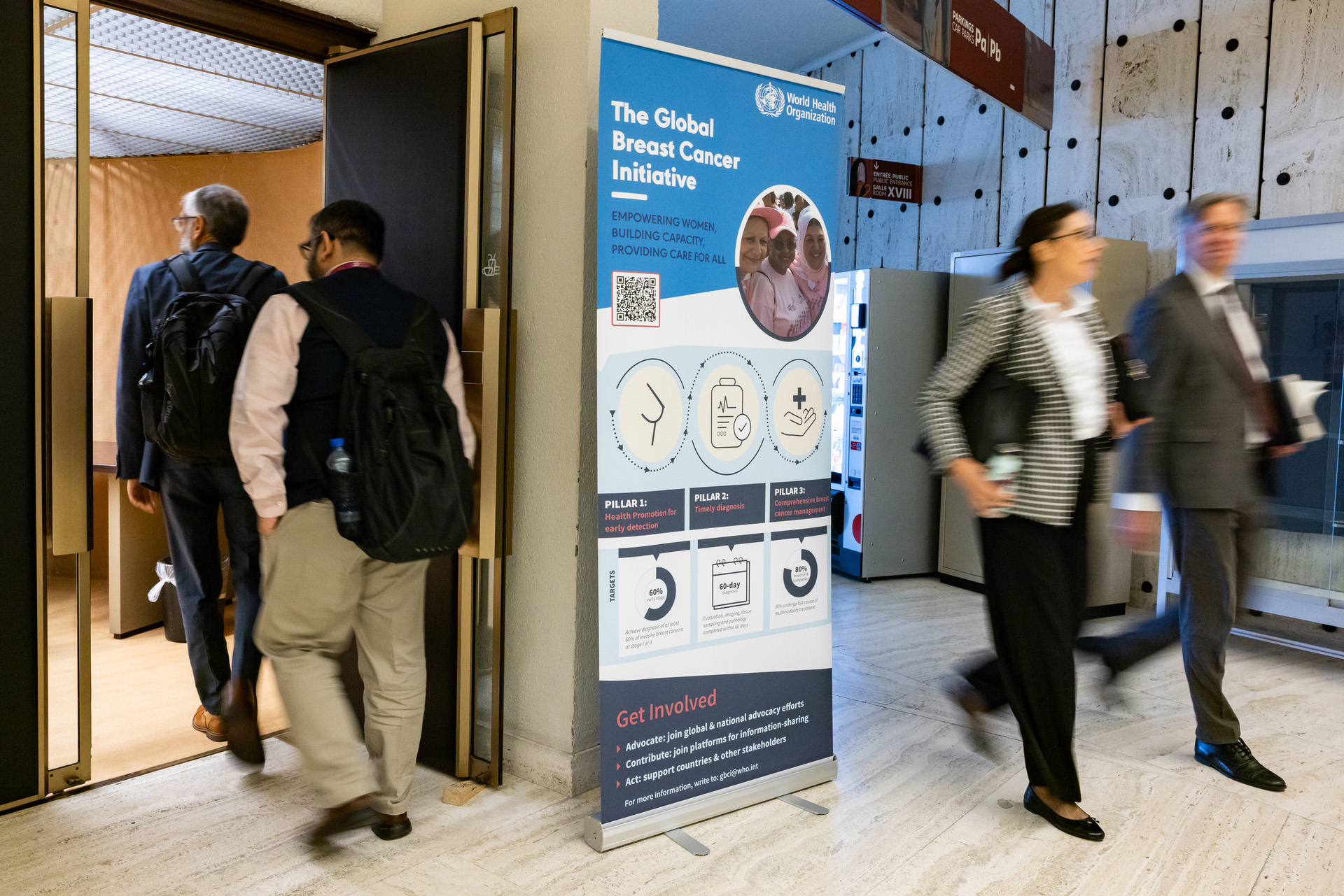 The backs of two people walking through a door and two more walking past a poster
