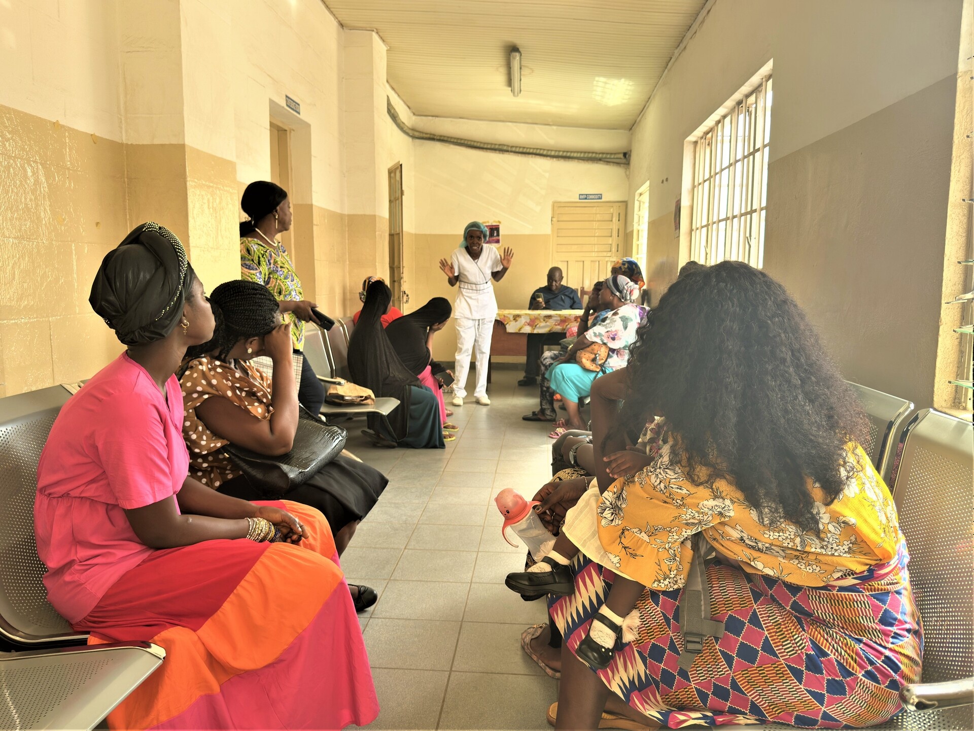 A health worker is talking to a group of female patients in a hospital corridor a man is behind a desk in the background