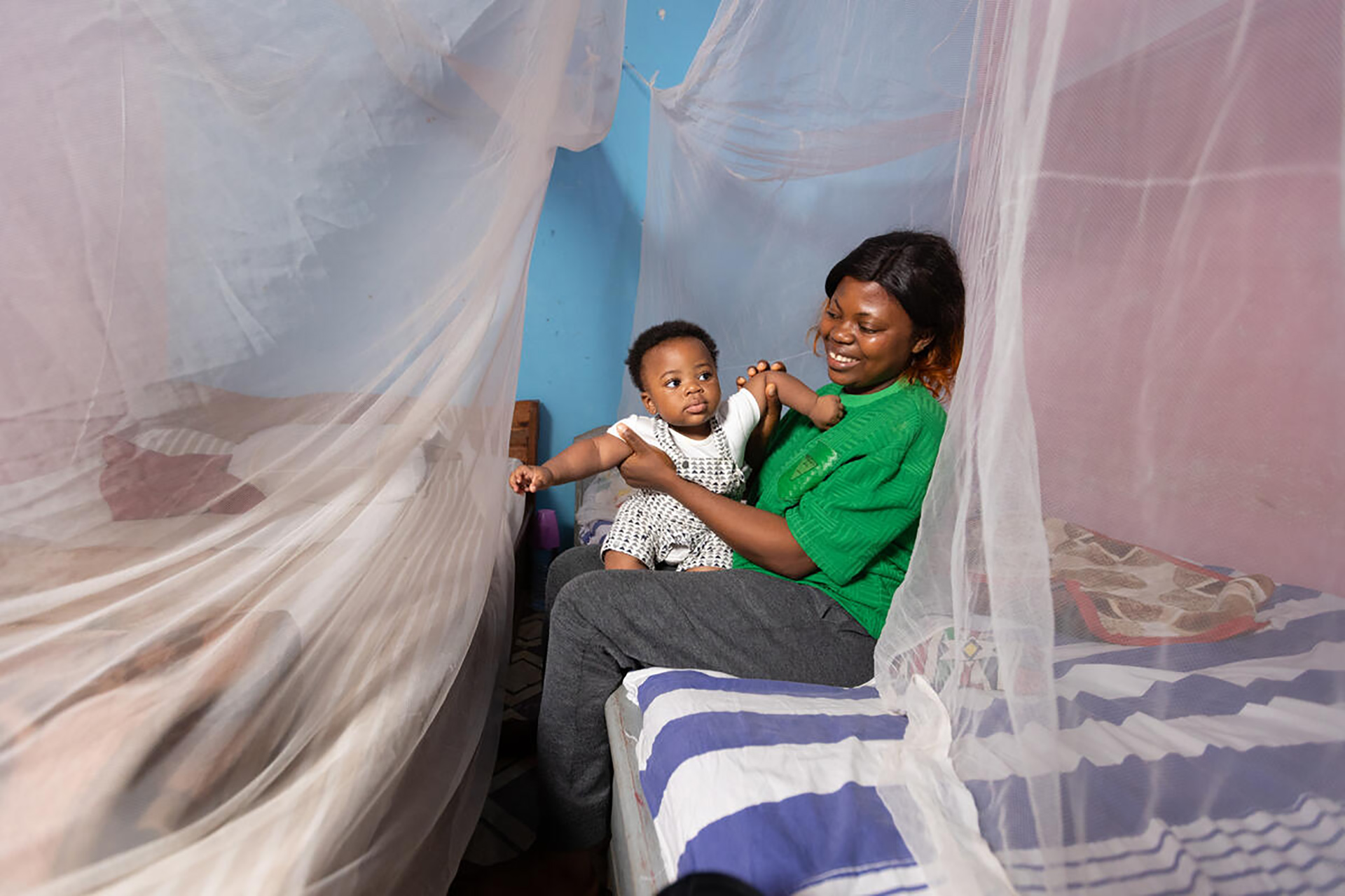 Smiling mother and her baby boy sitting together on a bed under a mosquito net in Cameroon