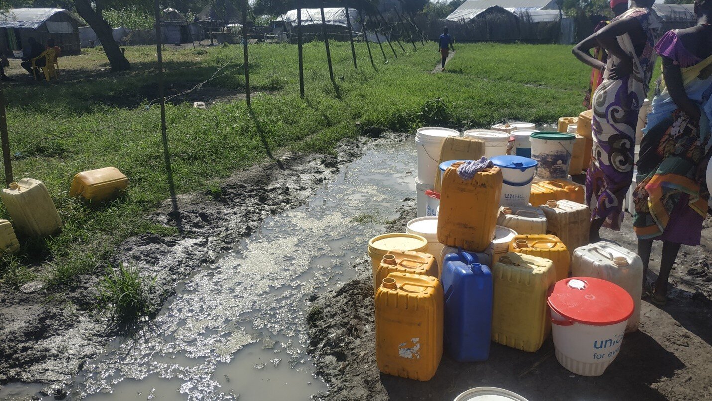 In a rural setting, next to a small pond of water, there are plastic containers in different shapes and colours. In the right corner of the image some women silhouettes are visible.