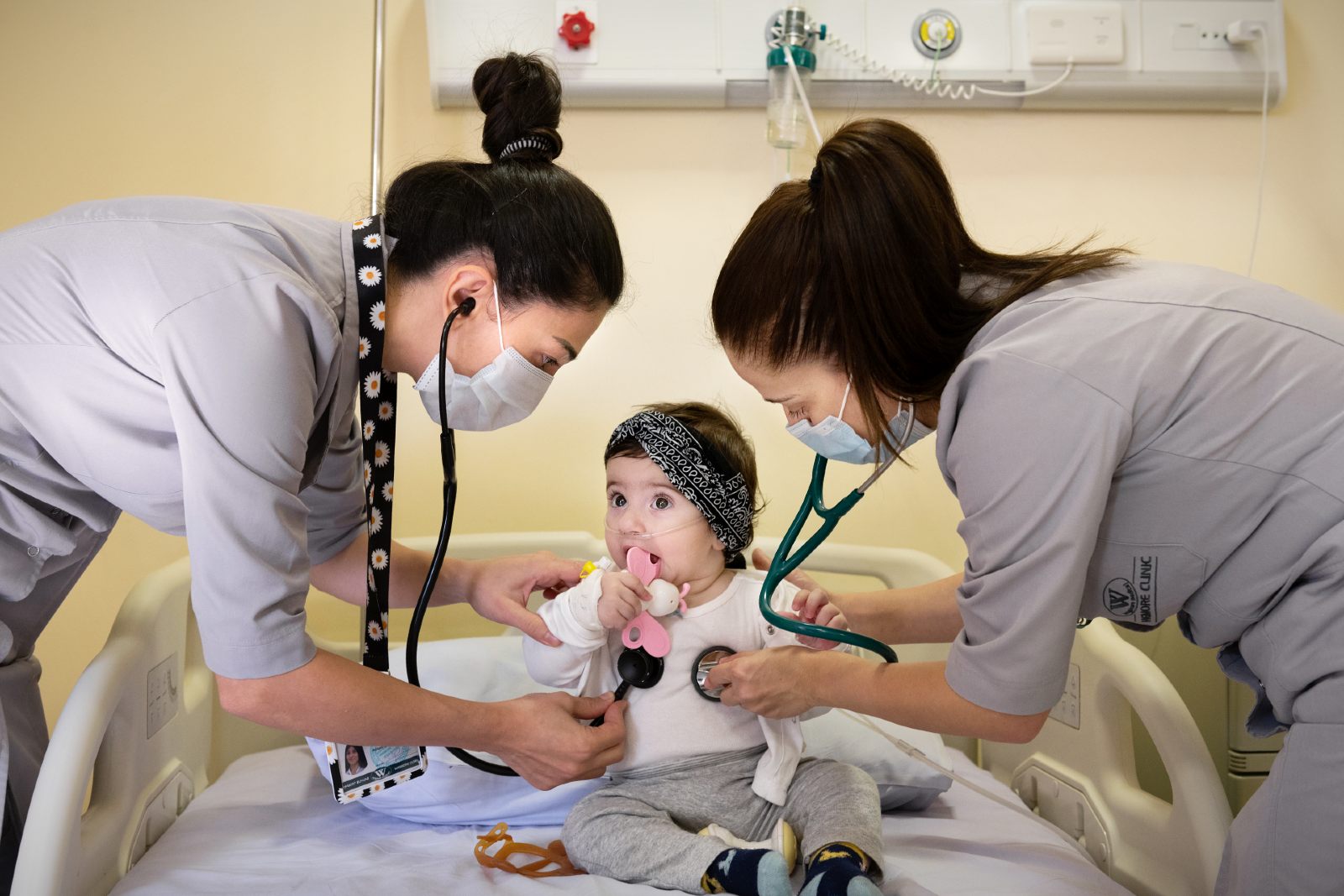 Two female pediatricians examine the lungs of a 9-month-old baby sitting in a hospital bed.