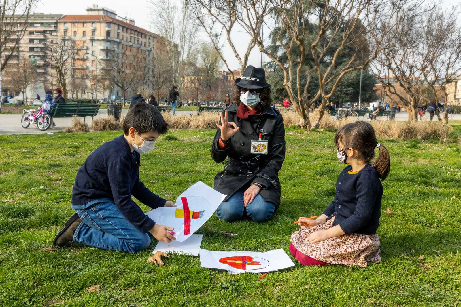 Two young children are sitting on the turf in a park together with a woman drawing anti-tobacco messages.