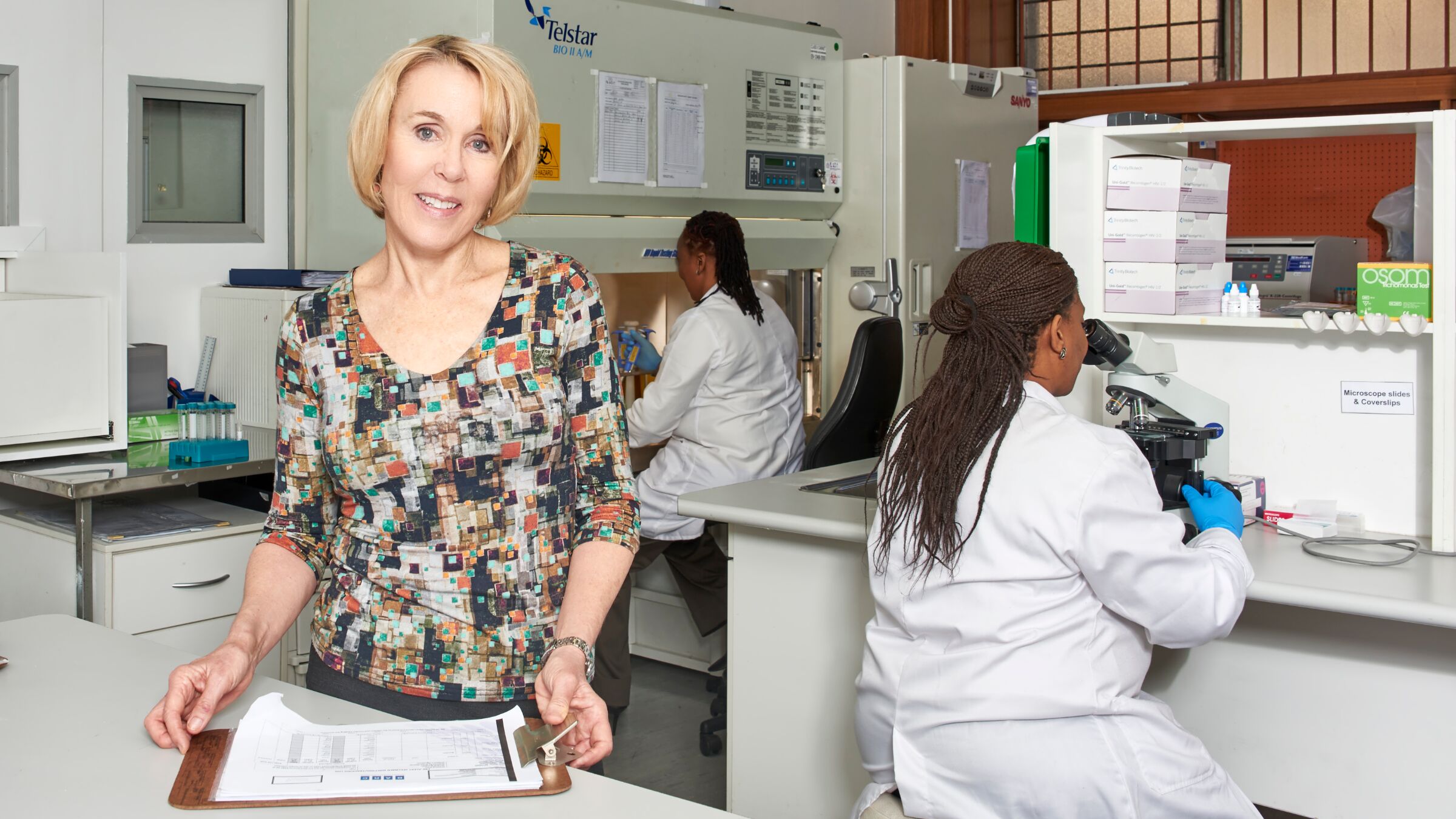 A woman looking into the camera is standing in a laboratory with two lab technicians in the background.