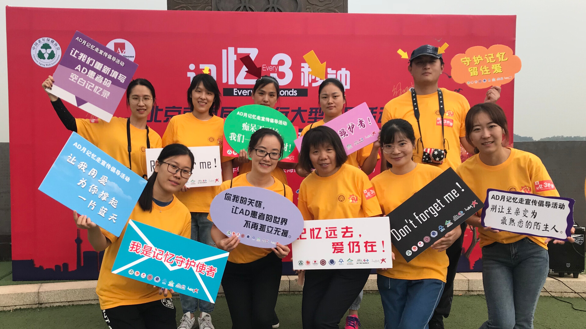 A group of people, all dressed in orange t-shirts, stands in front of a big backdrop holding up signs.