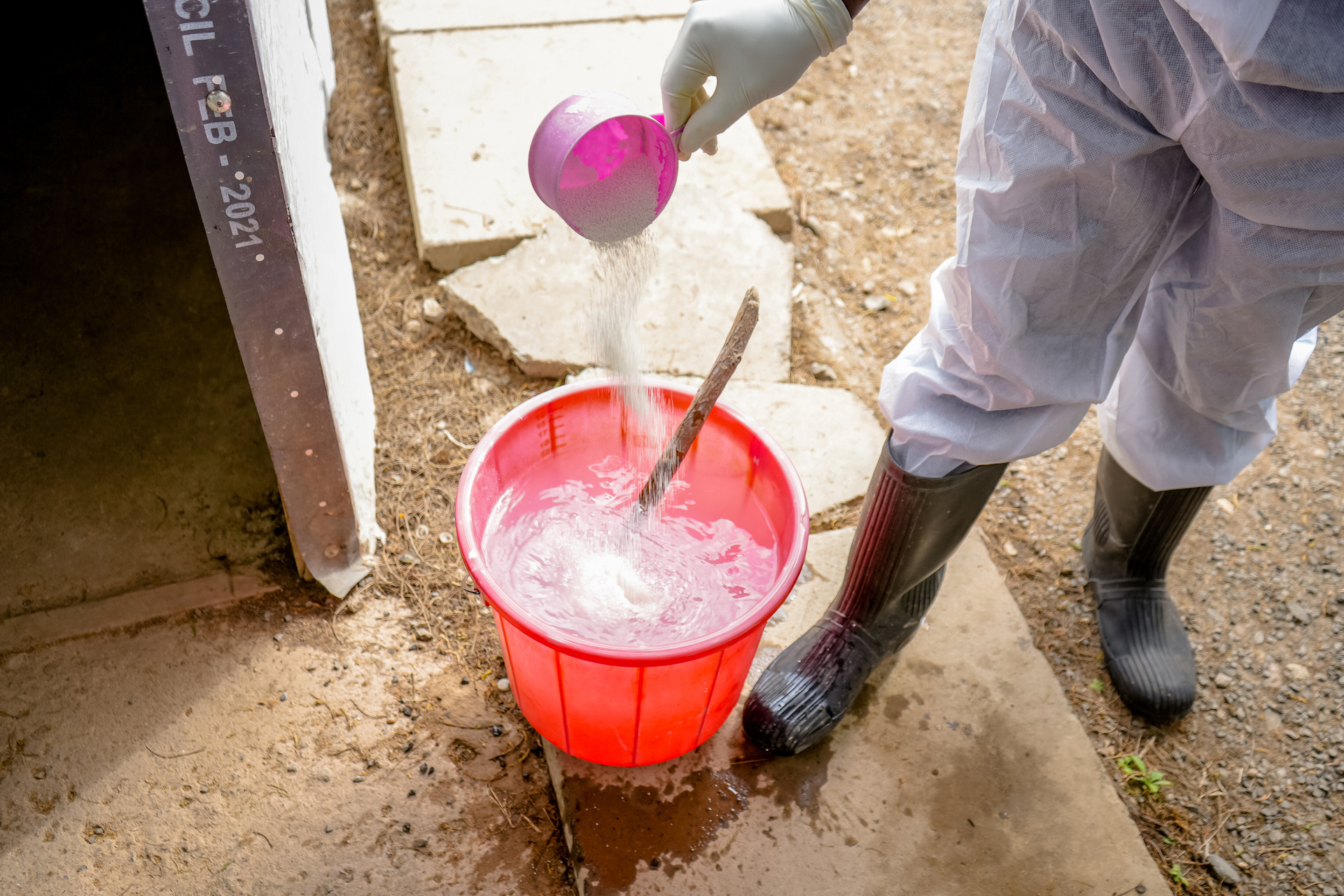 person in overalls and boots pouring chlorine into a bucket of water to be used to clean floors