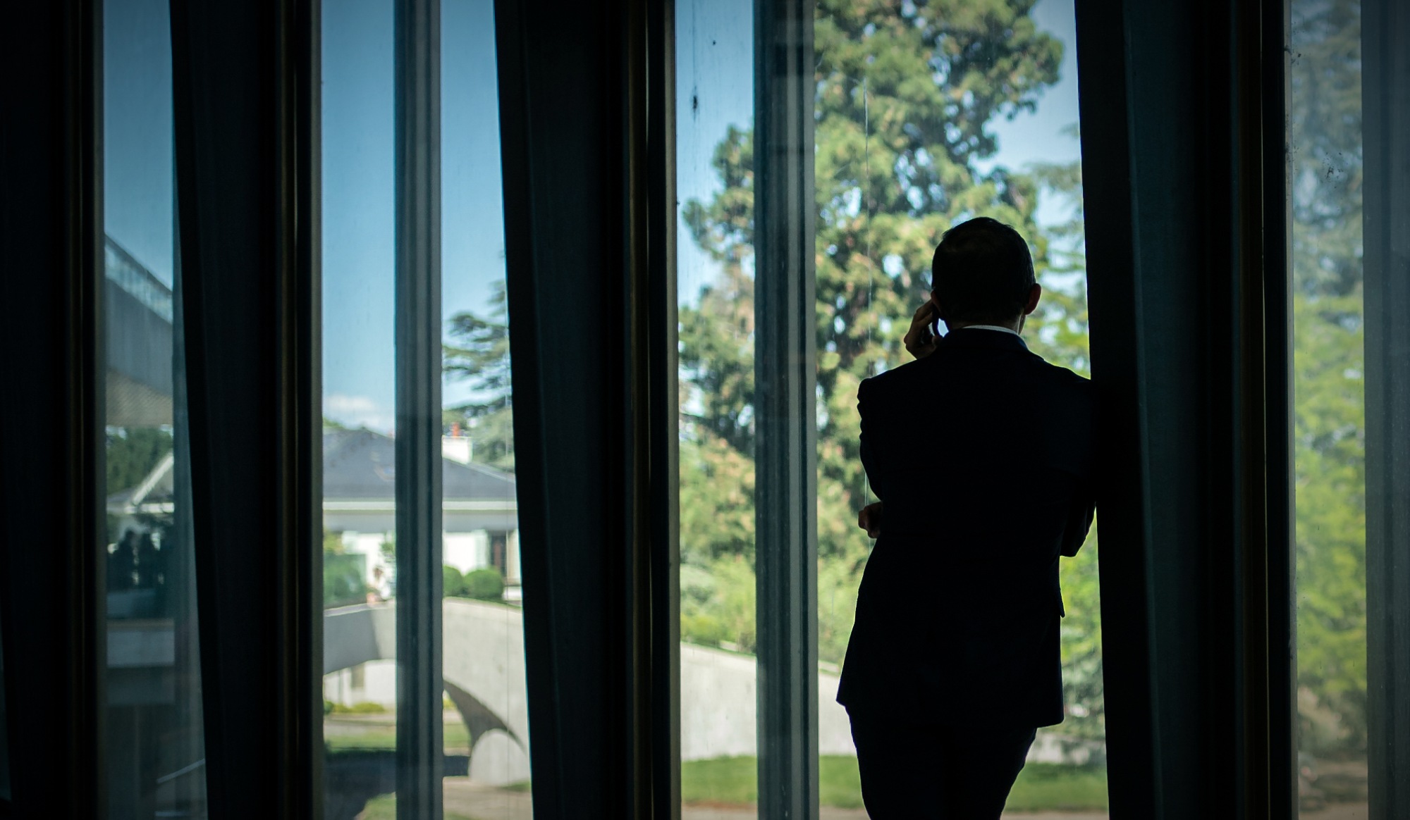 Reporting A silhouette of a man leaning against a pillar and talking on the phone, seen from behind and at a distance.