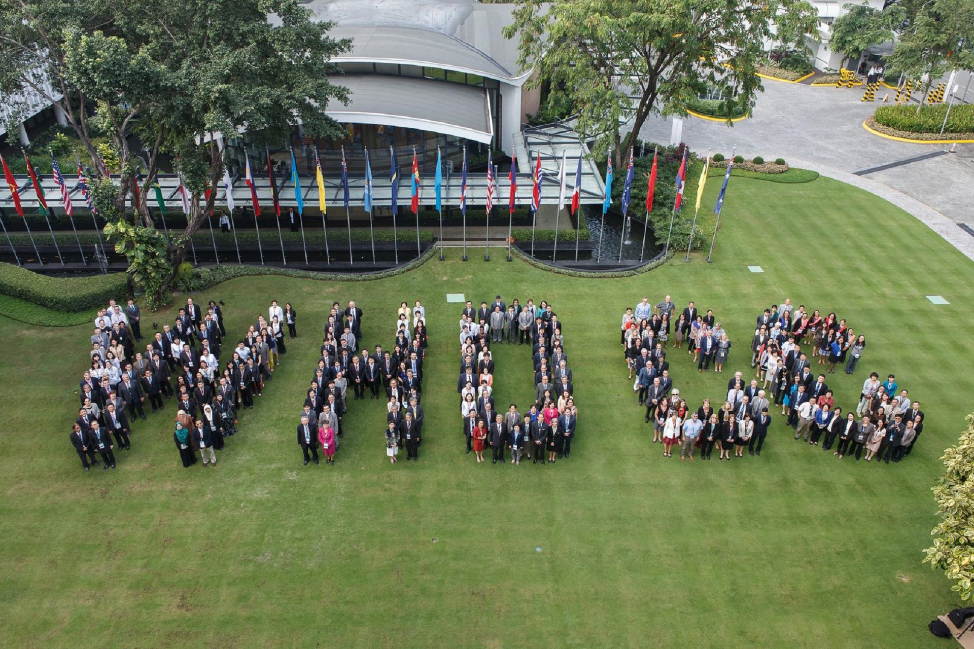 WHO Collaborating Centres Forum WHO Western Pacific A large group of people standing on grass, arranged to spell out the letters "WHO" in front of a building lined with international flags