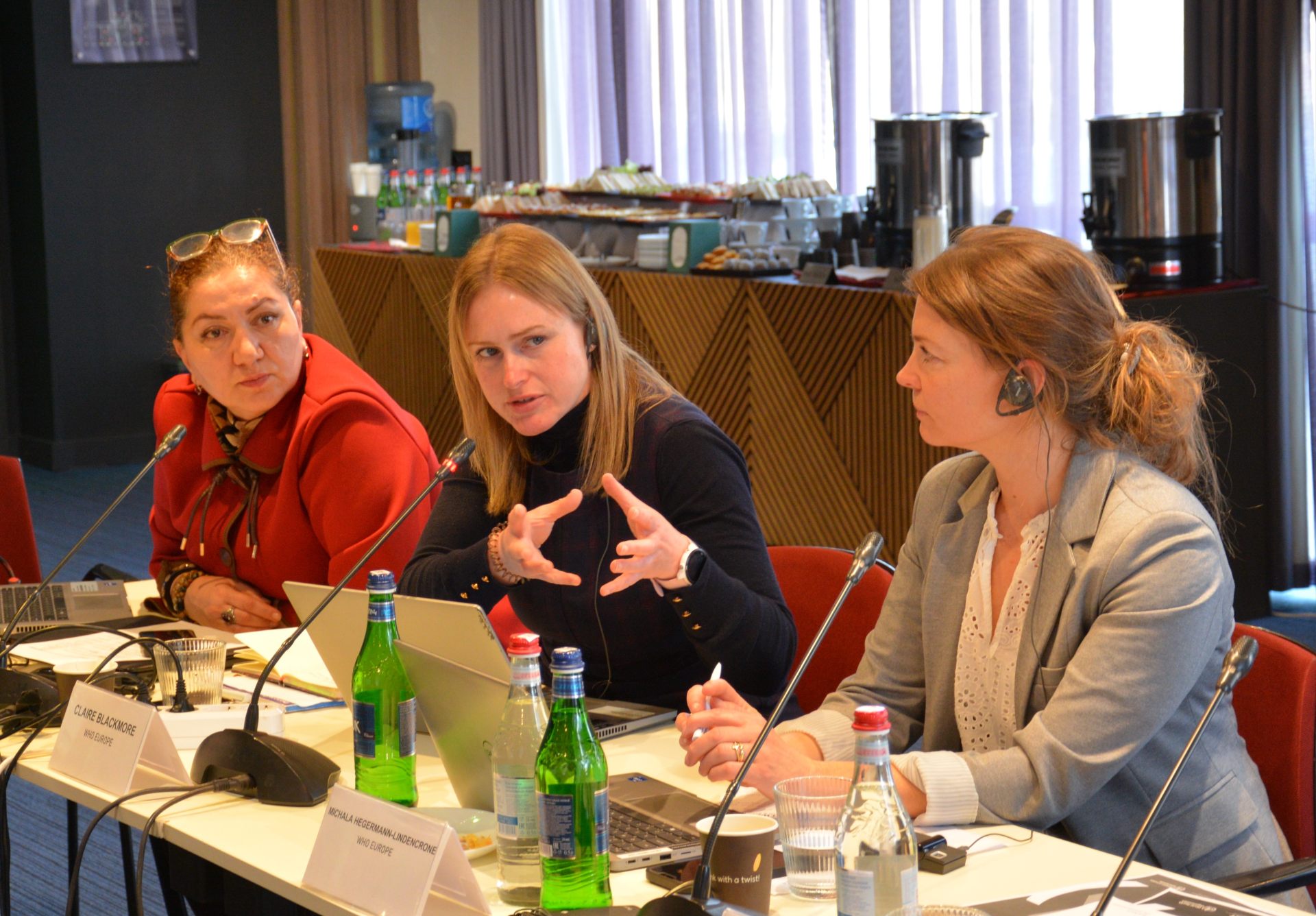 A speaker gestures while talking during a meeting, with two other women listening attentively